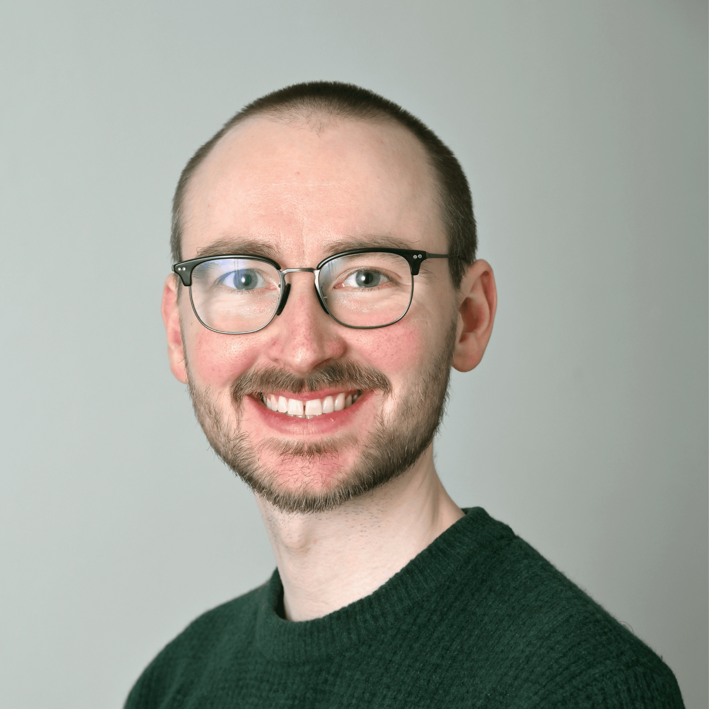 Colour photo of a white male with short hair wearing metal glasses and a dark green jumper.