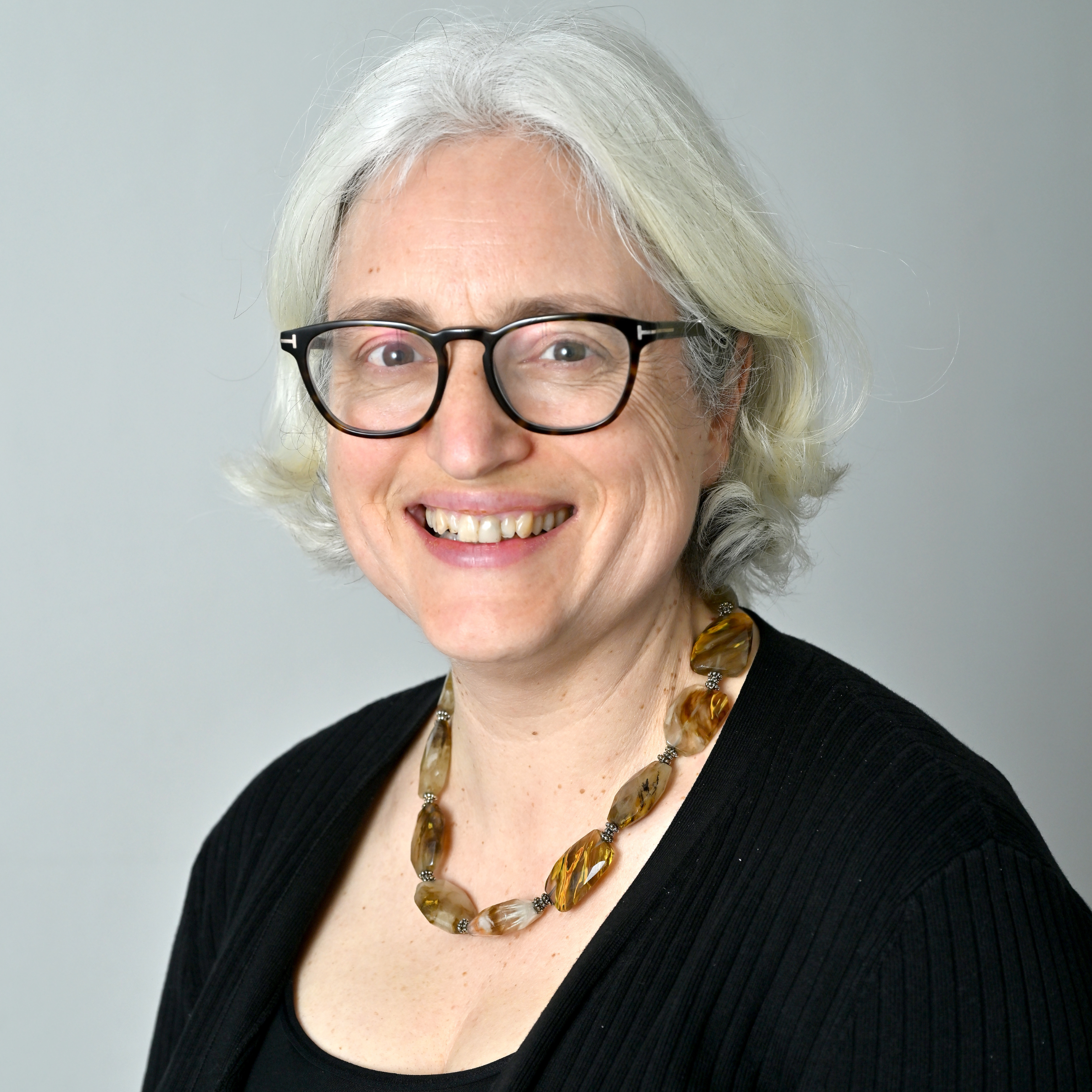 Head shot of a white middle age woman with short white hair and black glasses, wearing a black top and necklace