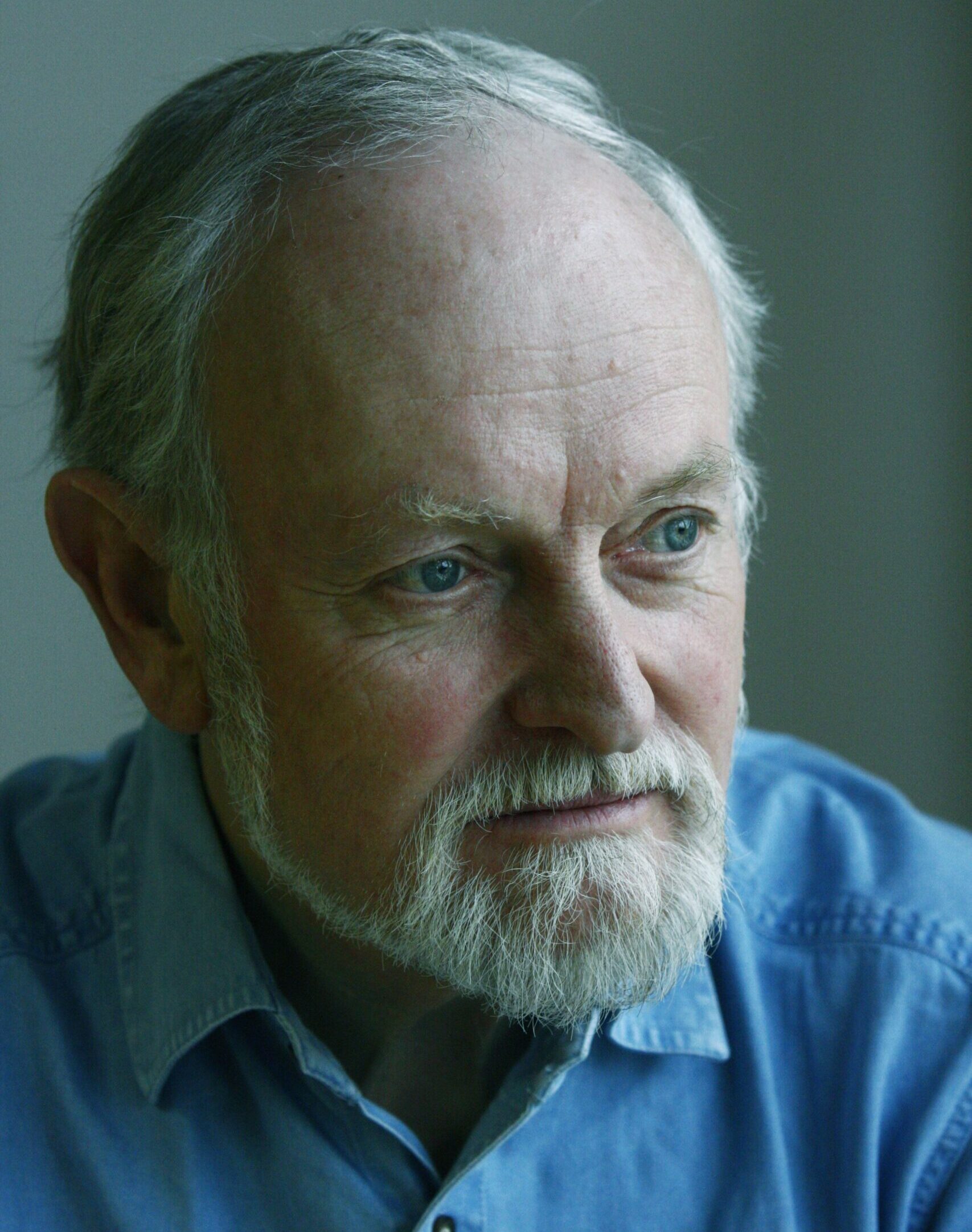 Colour headshot of a man with a white beard and blue shirt looking to one side