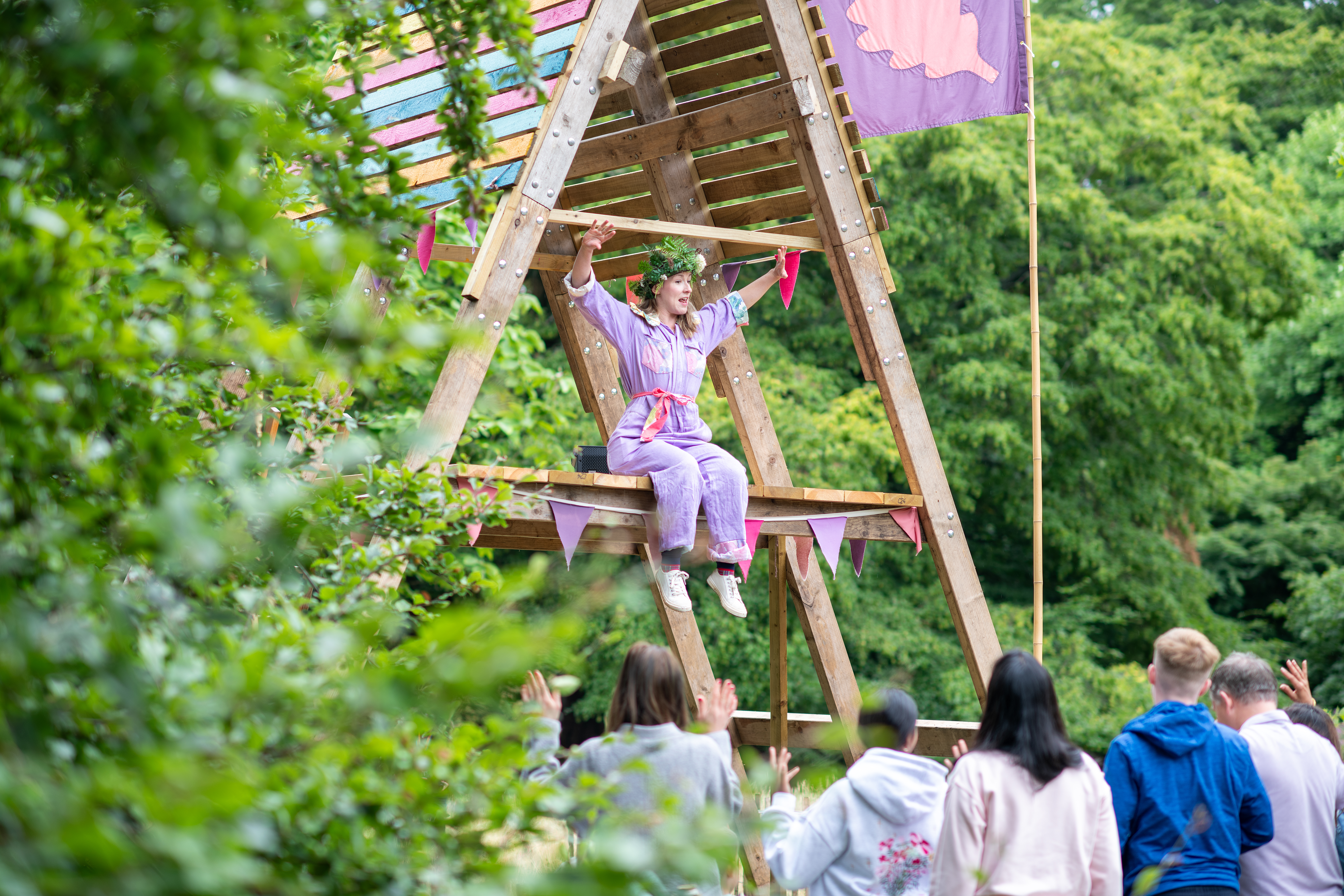A group of people standing in a woodland setting facing a large wooden triangular structure with someone seated on it