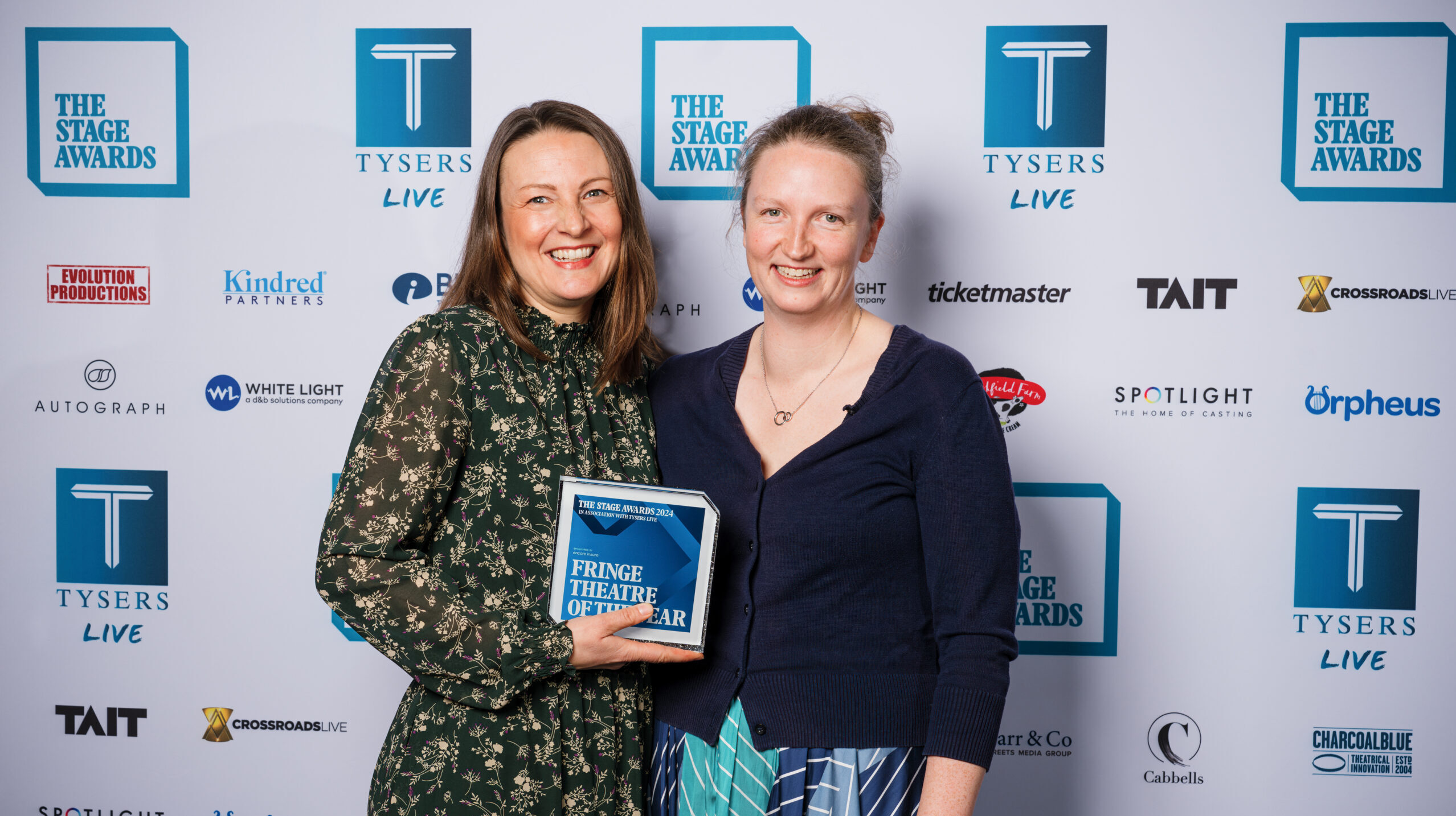 Two women holding an award staring before a Stage Awards banner logo board
