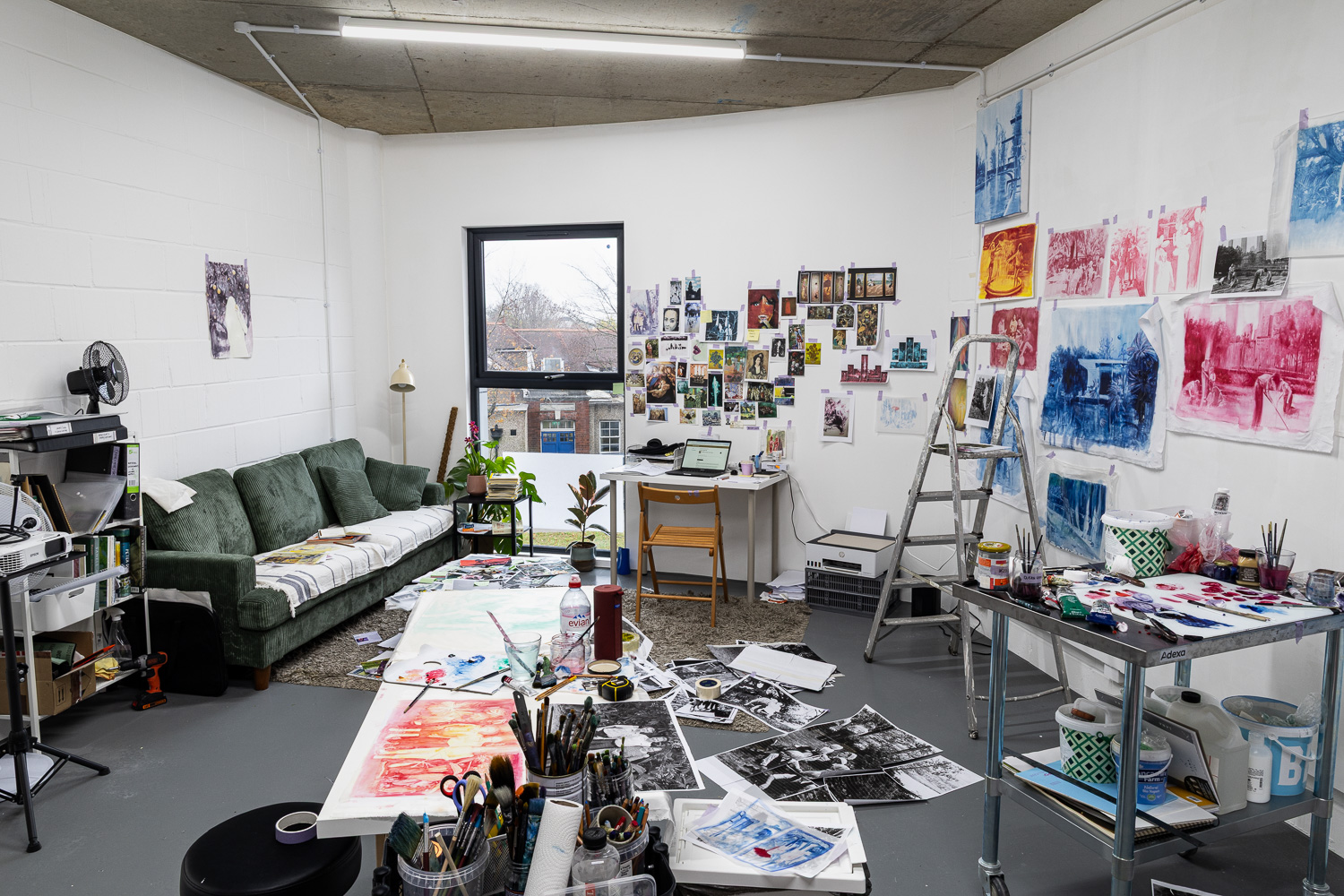 Colour photograph of an artist's studio with sofa, stepladder and walls and floor covered with bright artworks