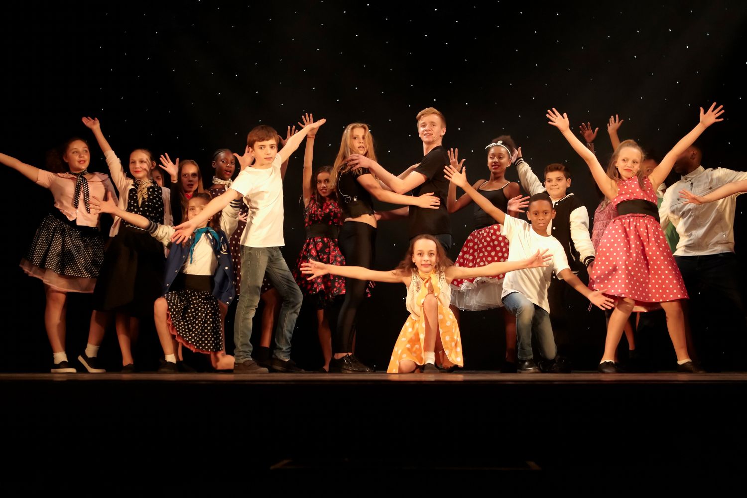 A group of children and young adults pose on a stage with arms extended.