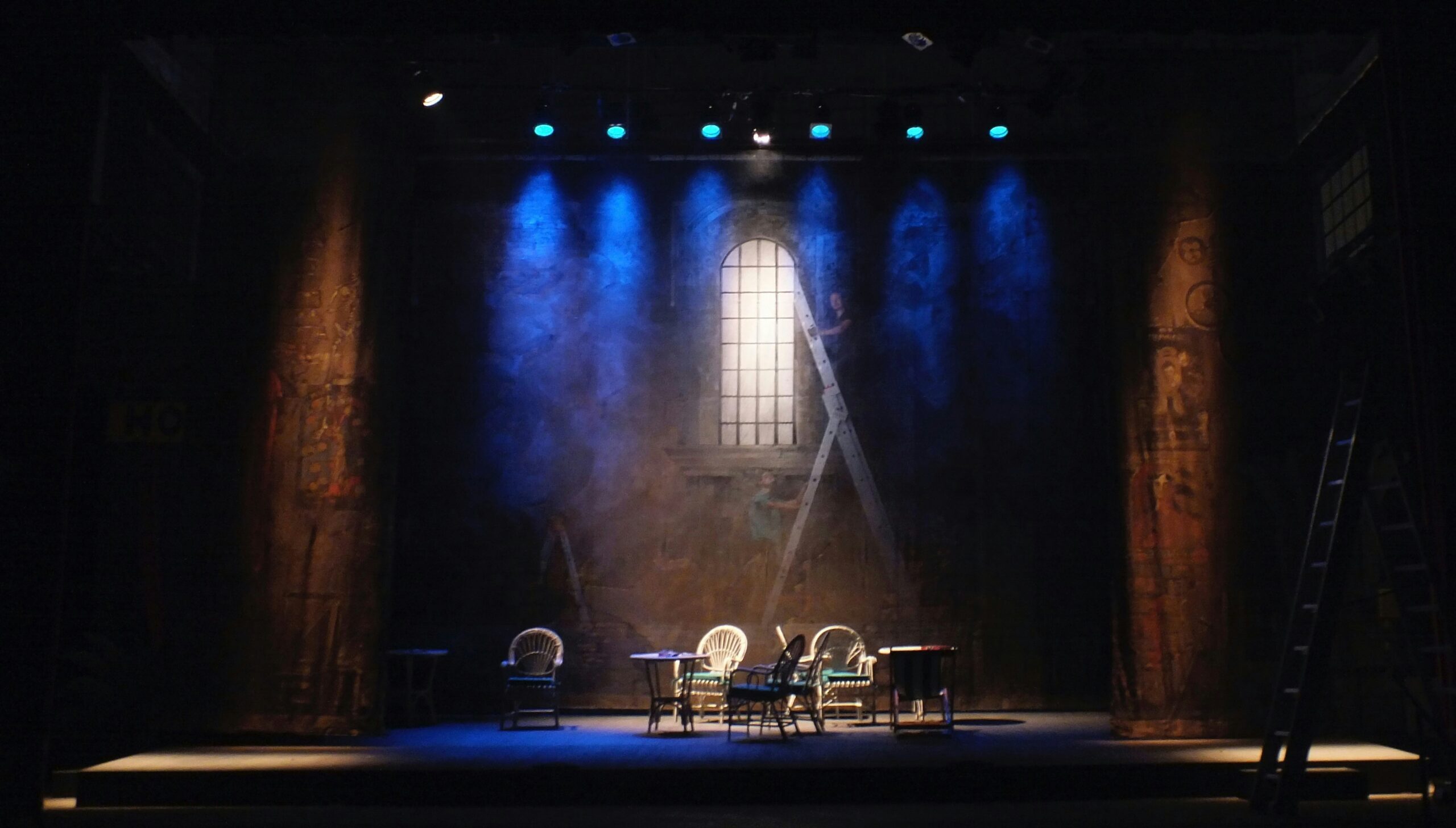 A theatre stage with chairs on the stage and a ladder behind with two people climbing it.