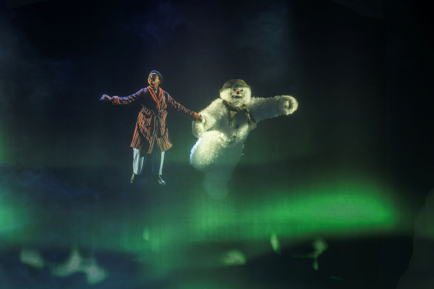 A production image from The Snowman. The Snowman and The Boy fly through the air above a green aurora.